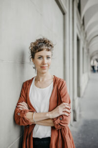 Evelyne Baumberger, trägt weisses T-Shirt und rostrote Jacke, lächelt in Kamera, steht mit verschränkten Armen in einem Säulengang